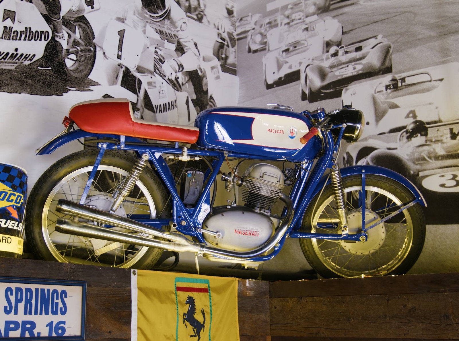 Vintage Maserati racing motorcycle on display against a wall of black-and-white racing photography at Turn 12